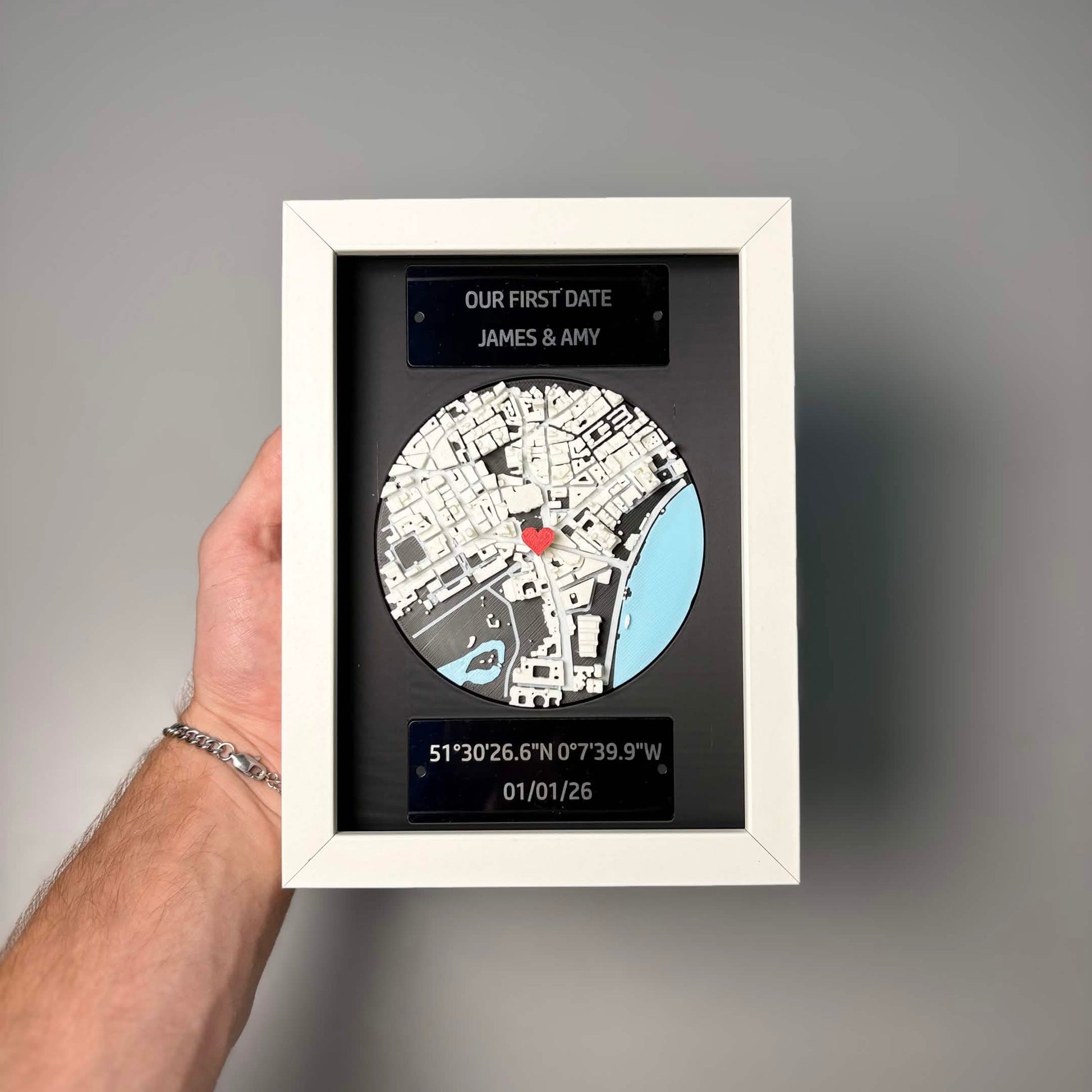 Personalised 3D map frame held in hand, showing scale, white frame, engraved plaques, and red heart location pin.