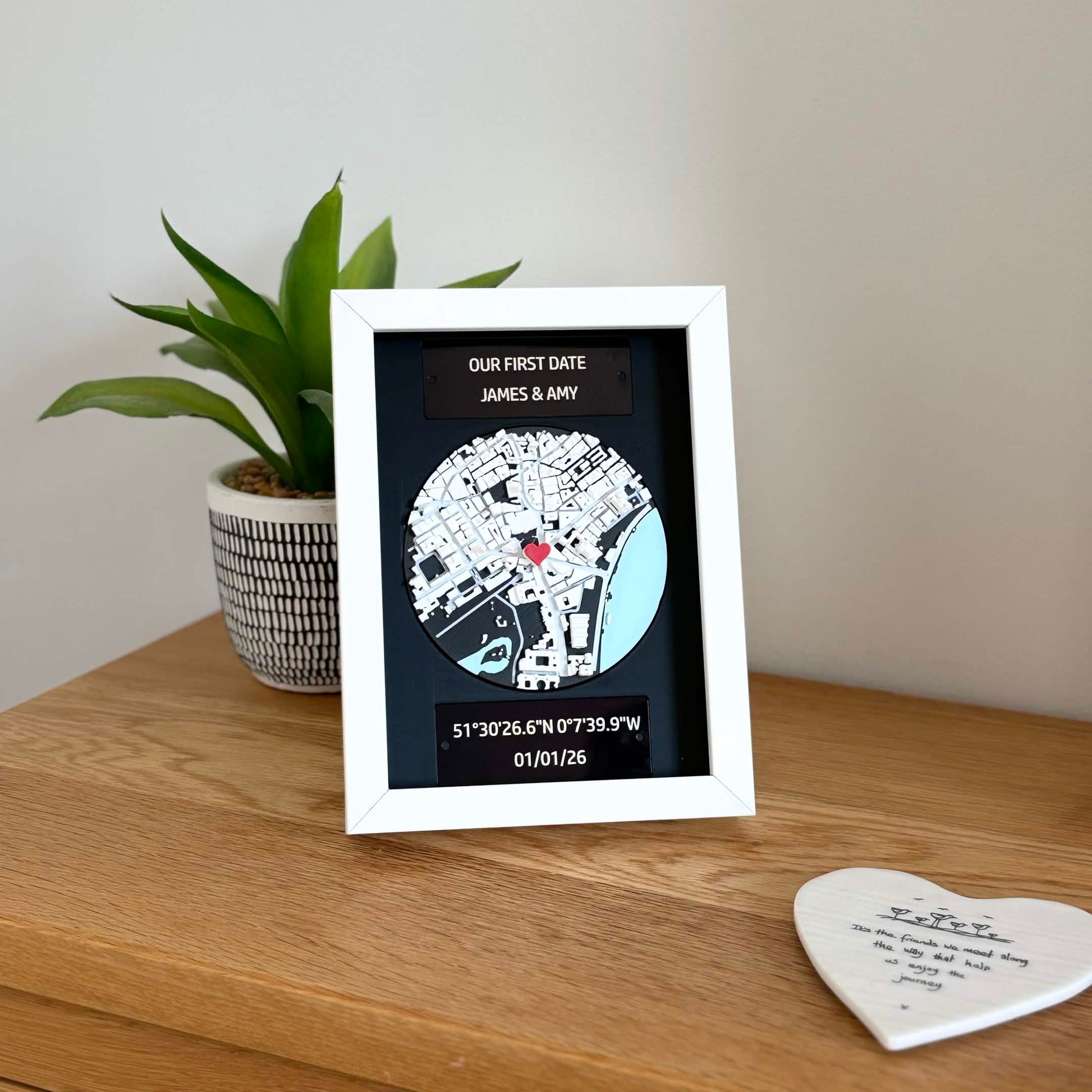 Personalised 3D map frame displayed on a wooden shelf beside a plant, engraved plaques and red heart pin visible.