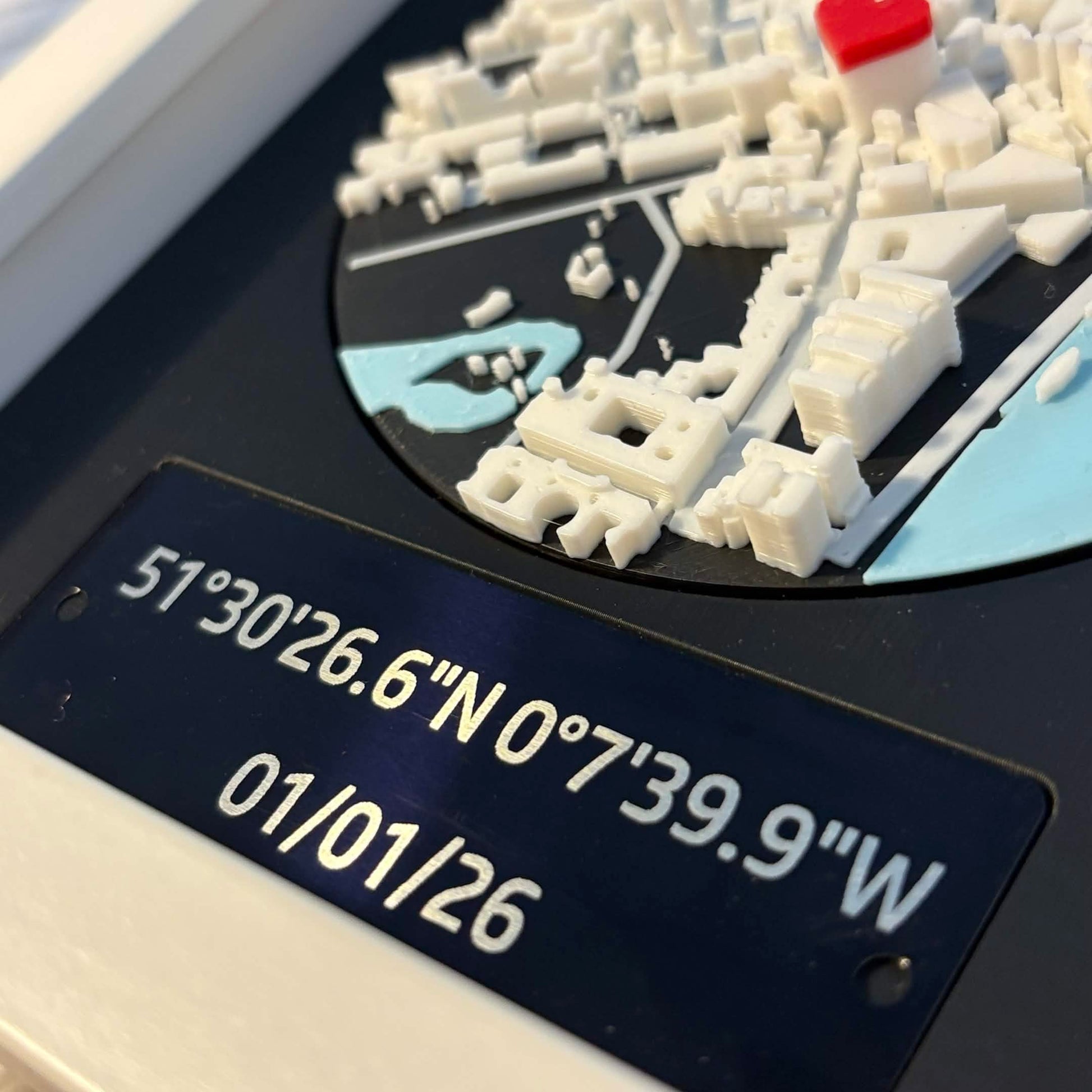 Macro shot of laser-etched aluminium plaque showing coordinates and date, with 3D printed map detail above.