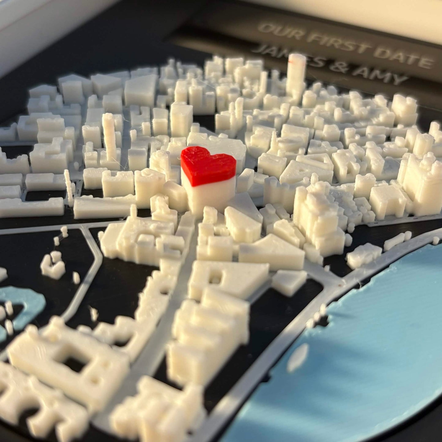 Macro close-up of a 3D-printed city map with red heart pin marking a custom location, engraved plaque behind.